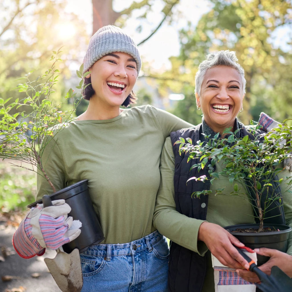 Two joyful friends hugging outdoors, holding potted plants and gardening tools.