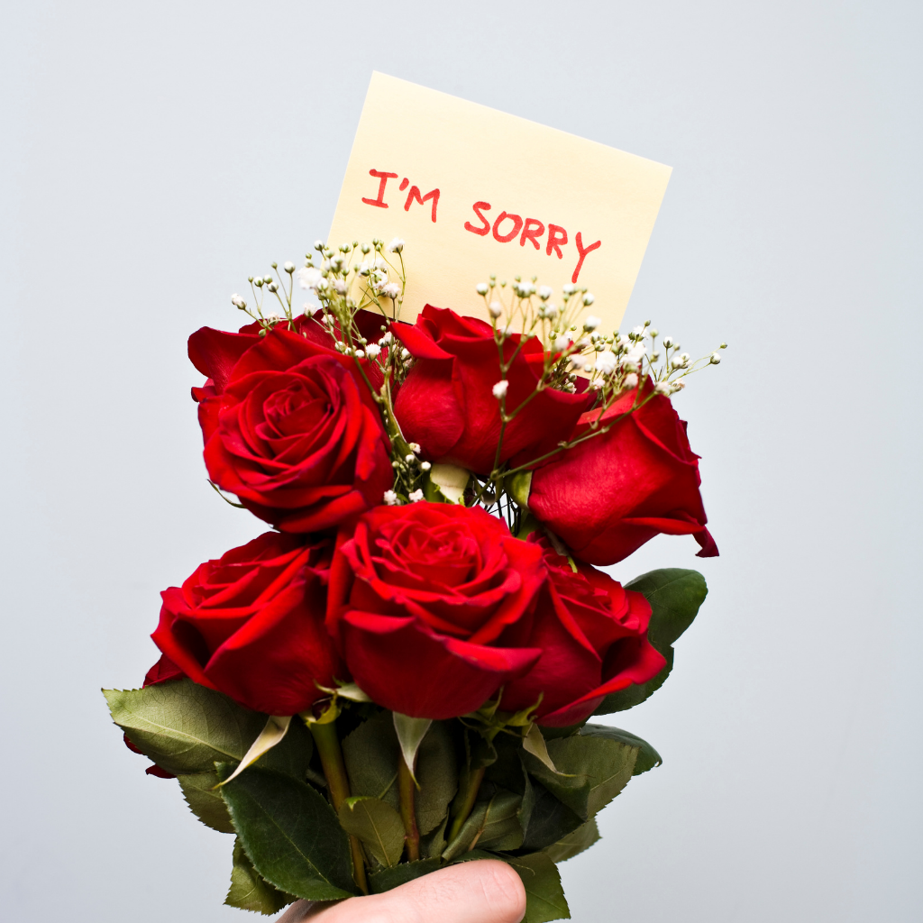 A hand holds a bouquet of red roses with a note saying "I'm sorry." The gesture expresses remorse and a desire for forgiveness.