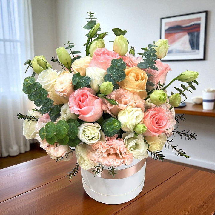 A floral arrangement with pink roses, champagne roses, white eustomas, and pink eustomas, placed in a white container with a pink ribbon.