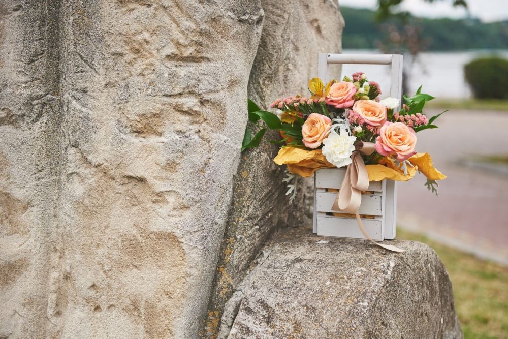 A charming arrangement of peach roses nestled in a rustic white wooden box, placed against an ancient stone ledge with a tranquil lake in the background, perfect as flower decor for outdoor weddings.