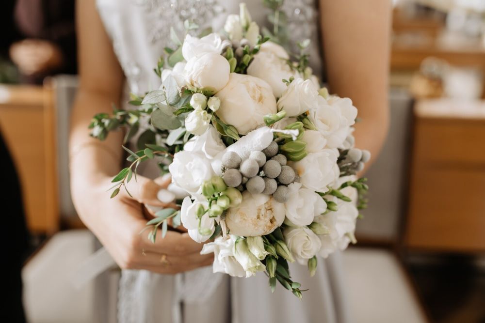 A stunning white wedding flower bouquet with white roses, peonies, and accents of green leaves, ideal for wedding centrepieces and bridal bouquets.