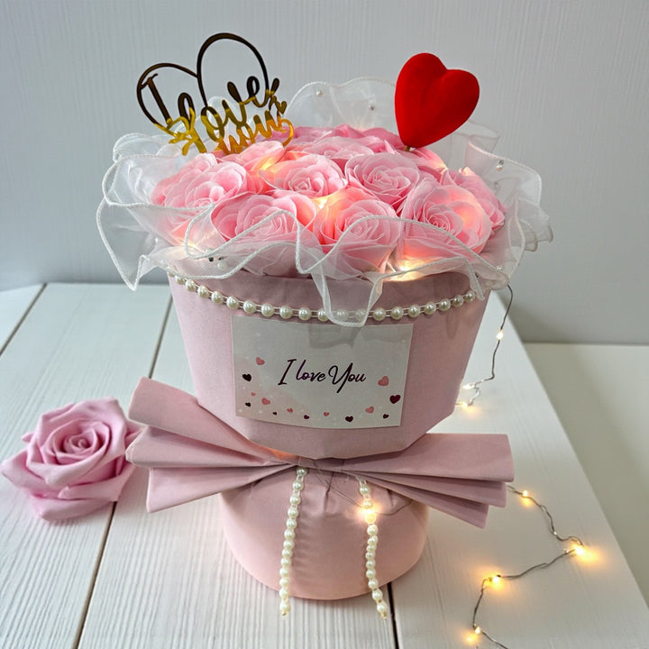 Decorative flower arrangement with pink roses, 'I Love You' sign, and fairy lights on a white surface.