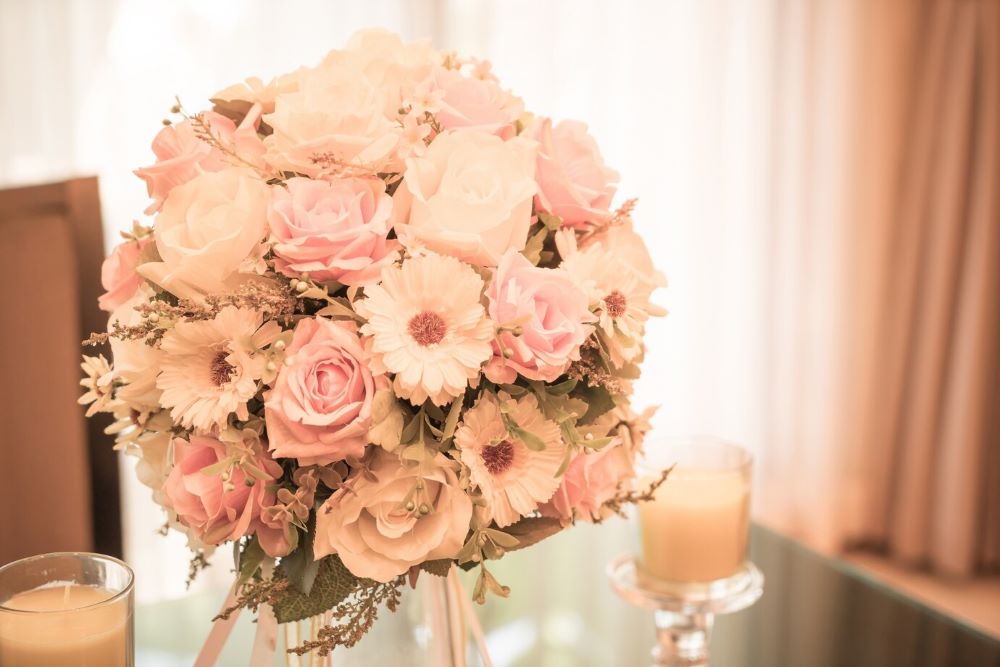 A luxurious wedding flower bouquet of peach roses, pink gerberas, and delicate white blossoms, elegantly arranged on a table beside a softly glowing candle.