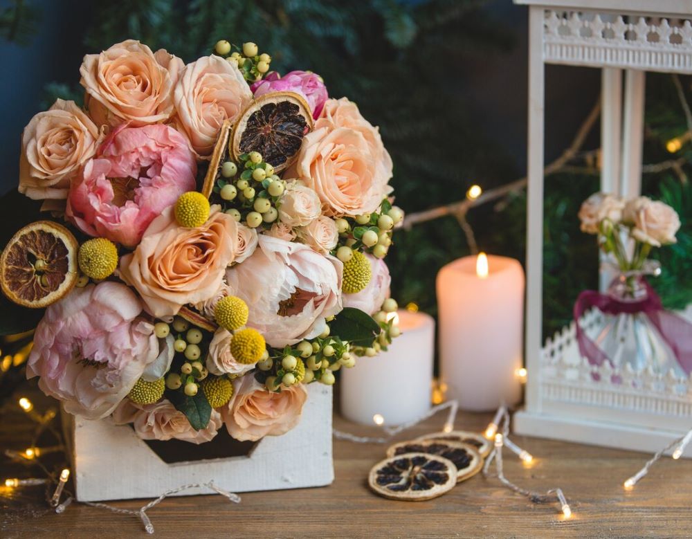 An elegant wedding table centrepiece featuring flowers like peach roses and pink peonies along with dried citrus slices, illuminated by candlelight, creating a romantic ambiance for a special evening.