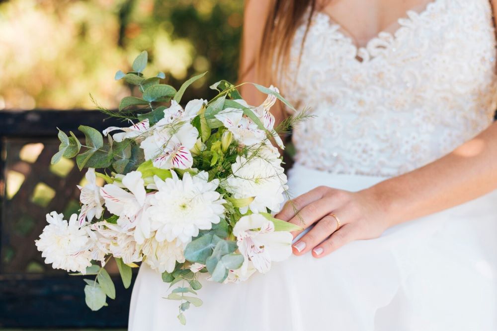 A bride in a beaded gown delicately holds a simple yet elegant white floral bouquet with alstroemeria and daisies.