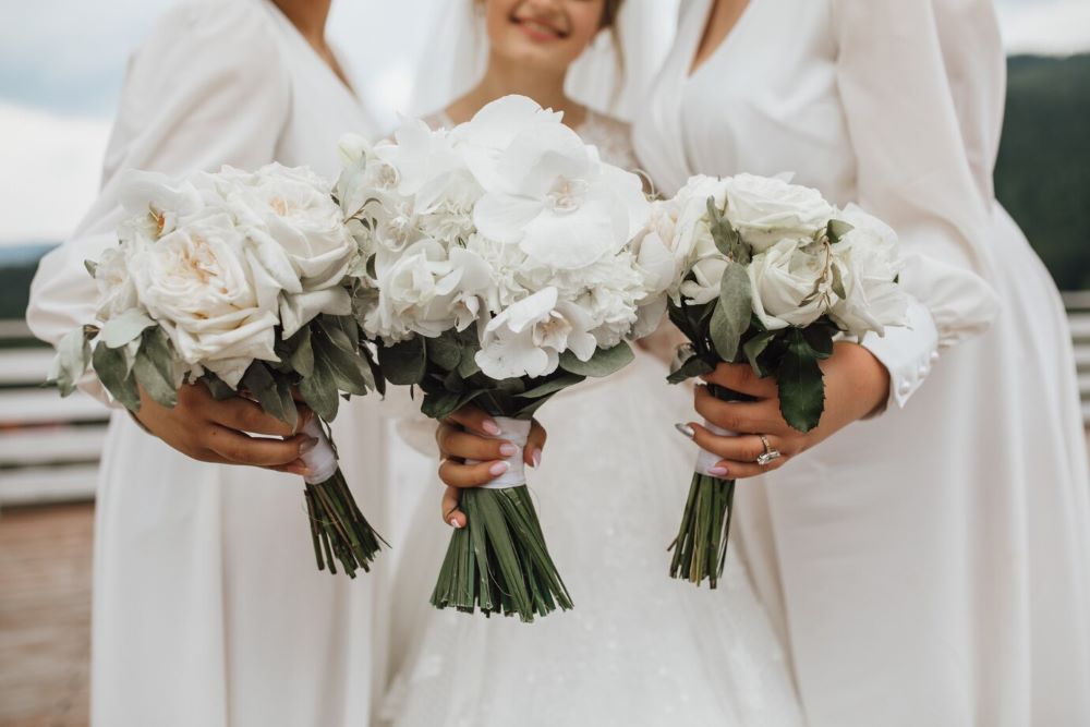 A bride and her bridesmaids in white dresses, each holding a beautiful white wedding bouquet, consisting of roses and orchids.